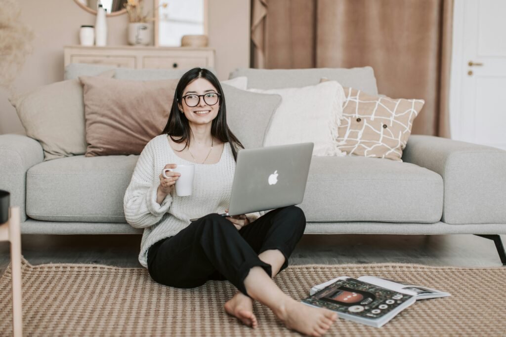 Full body happy young female in casual clothes sitting on floor near sofa with cup of tea and laptop while surfing Internet
