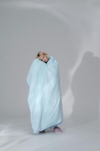 A woman standing wrapped in a blue blanket in a minimalist studio setting.