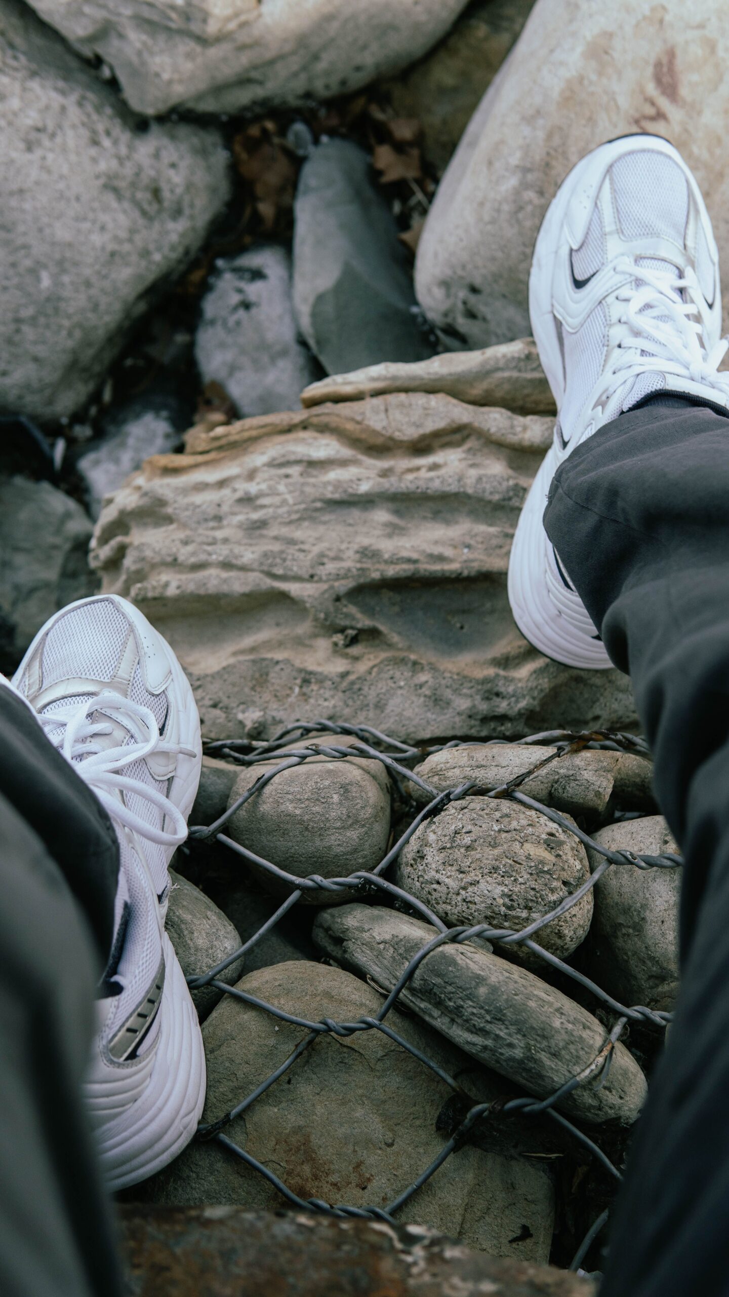 Person's legs and white sneakers over rocks and wire mesh outdoors.